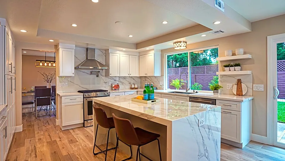 White shaker kitchen with a marble-look waterfall island, leather stools and a window over the sink
